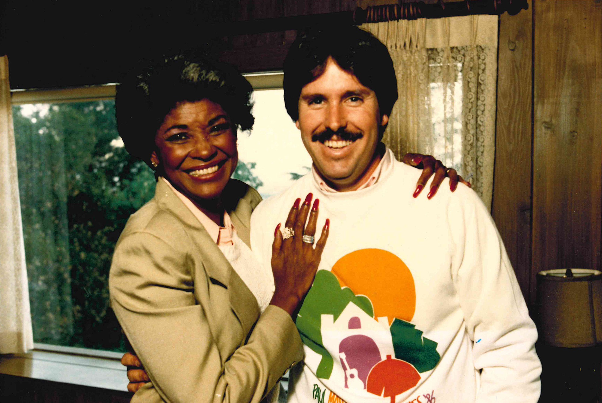 A white man in a mustache and colorful sweatshirt poses against a wood paneled wall with a striking Black woman in a tan suit jacket and decorative rings.