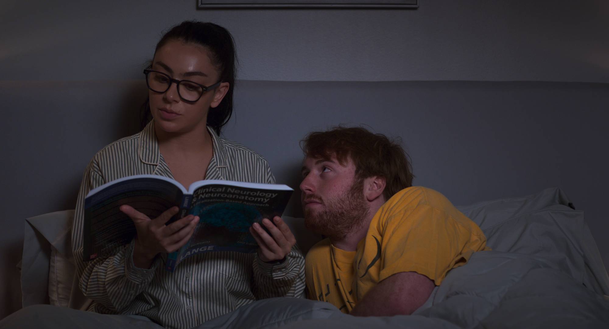 a woman wearing glasses reads a book in bed while her boyfriend looks at him