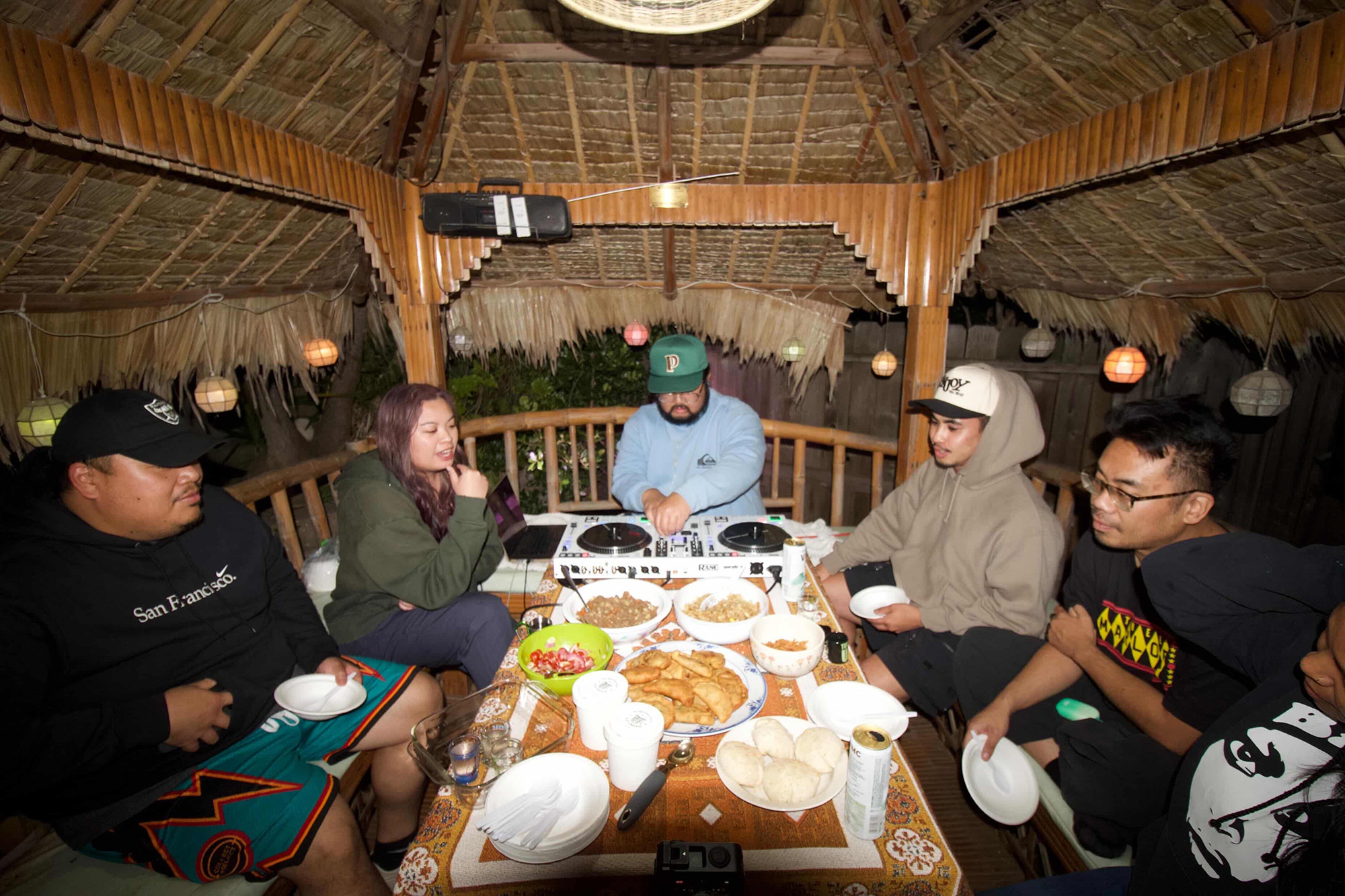 A DJ works the turntables inside a hut, surrounded by Filipinos digging into a spread of food.