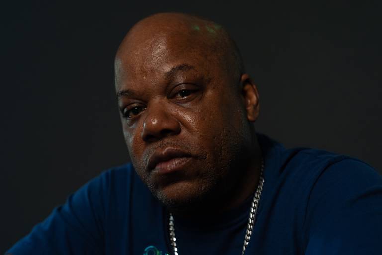A close-up portrait of a Black man, bald with a chain necklace and blue shirt, looking into the camera