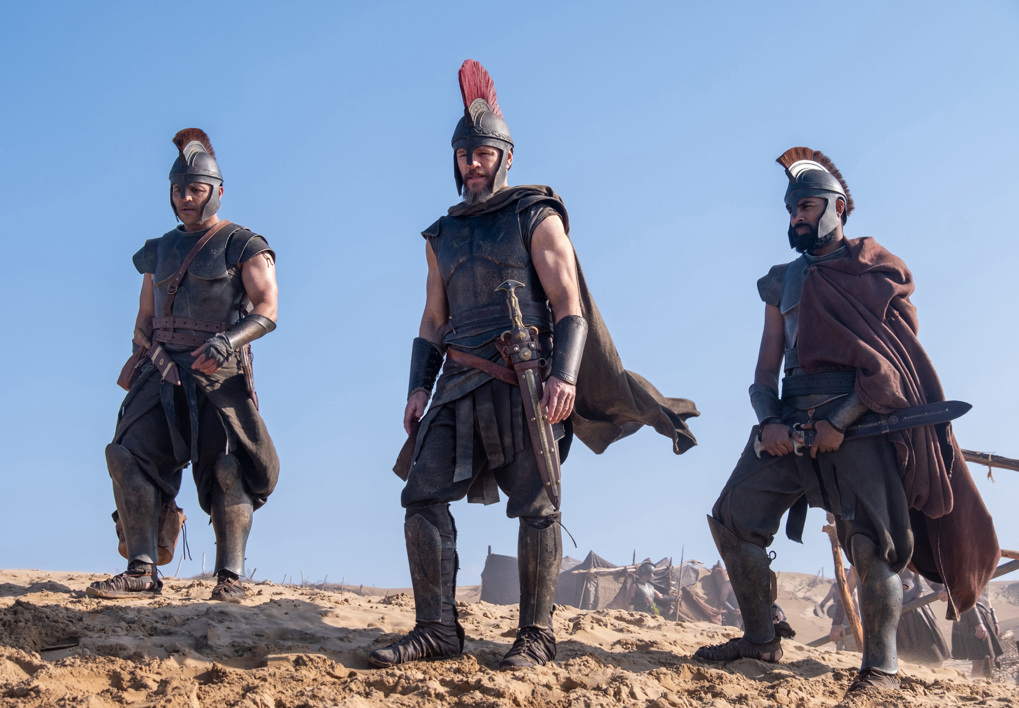 three men in sword and sandal attire stand on sandy hill