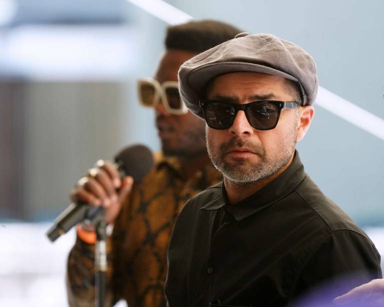 A man in Ray-Ban style sunglasses, black shirt and a newsboy cap looks to the right while a Black man in white sunglasses clutches a microphone in the background