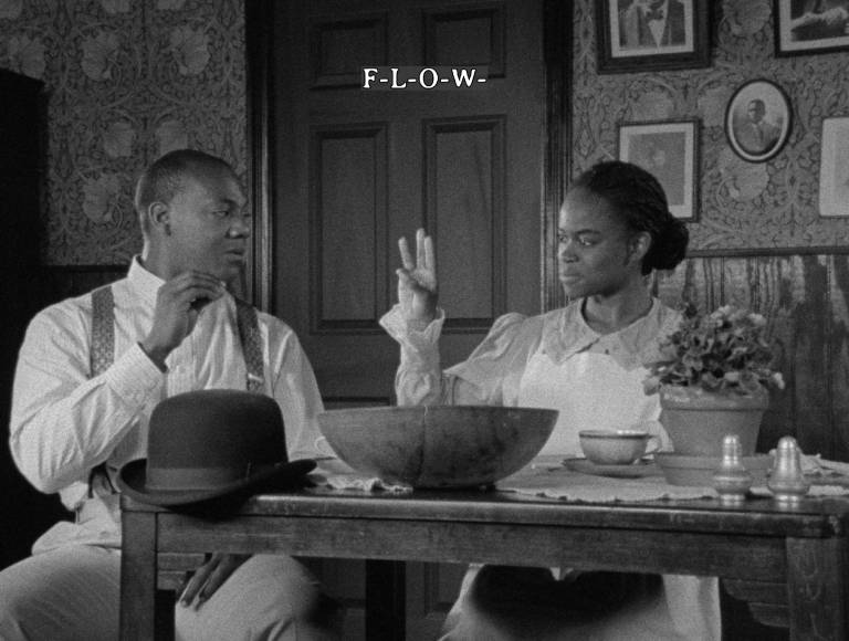 Black man and woman sit at table in 1910s dress, sign 'F-L-O-W' to each other in ASL