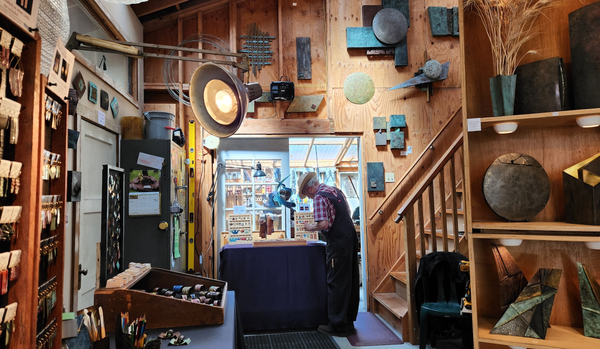 A white-haired man wearing coveralls and a checkered shirt stands looking closely at a table of displayed jewelry. He is inside a studio with wooden walls that are covered in abstract geometric art. Shelves of vases stand close to the camera, opposite a wall of neatly displayed earrings. To the right of the man are stairs leading up.