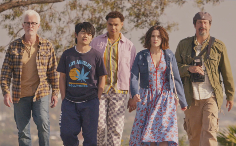 A white woman in a floral dress walks with purpose surrounded by four men of different ages and races dressed casually but all wearing serious expressions.