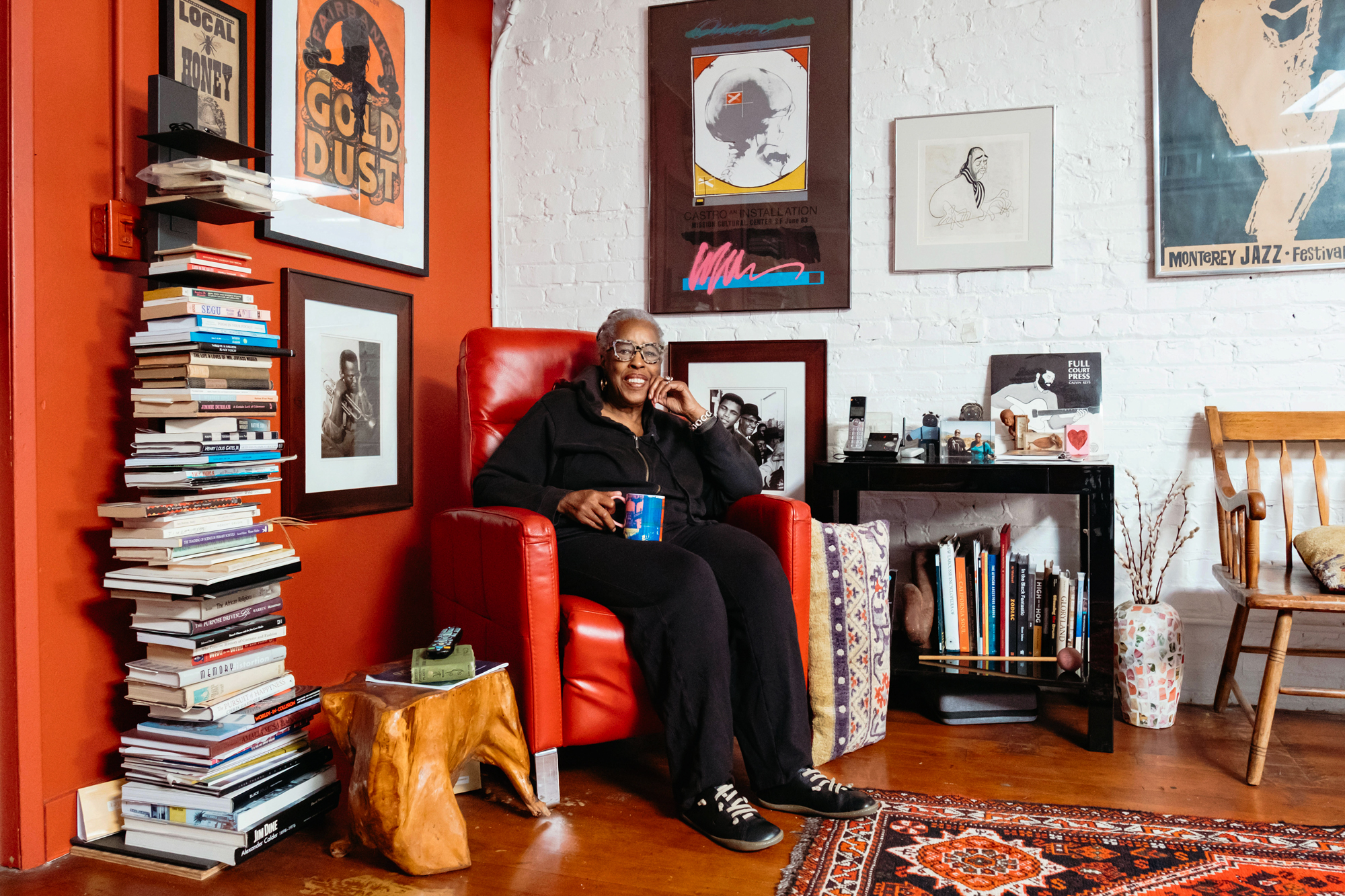 older Black woman smiles in armchair among books and framed artworks