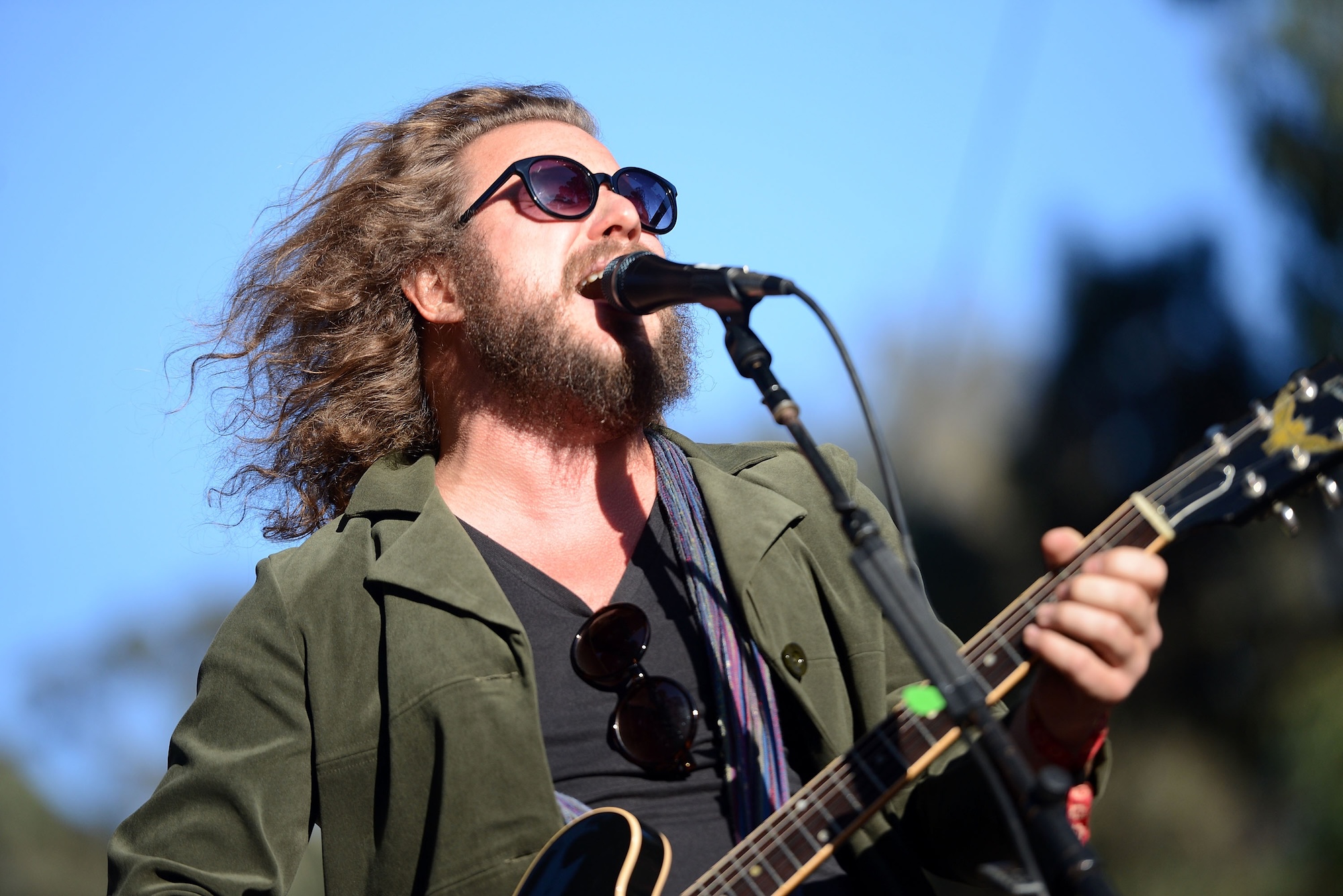 A white bearded man with shoulder length wavy hair sings into a microphone and plays electric guitar, outdoors. He is wearing a black t shirt and green jacket.