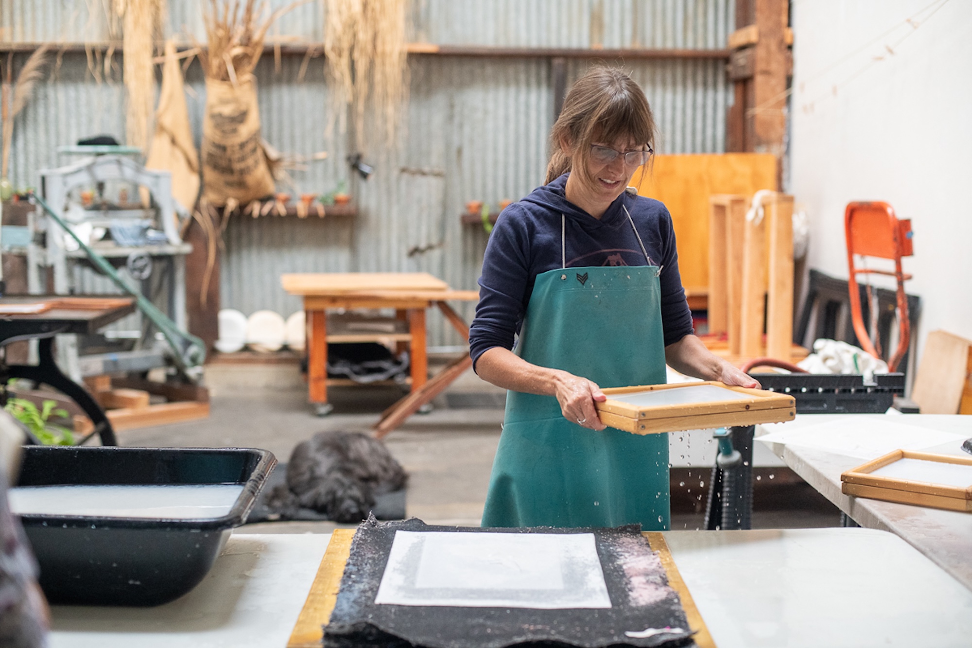 A woman wearing glasses and an apron, brown hair pulled back into a ponytail, holds a dripping wet wooden rectangular box in front of her. Behind her is a corrugated iron wall, multiple work tables and a black shaggy dog.