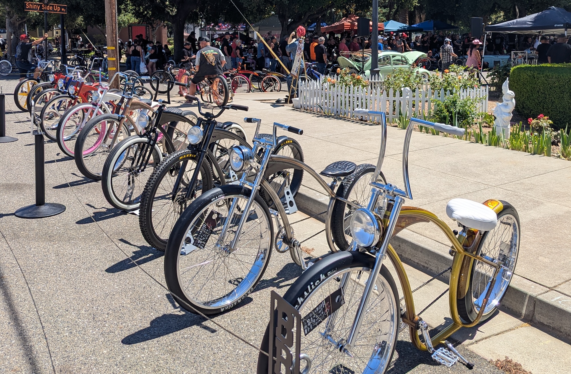 A row of custom-made colorful cruiser bicycles, lined up in front of a sidewalk. A party and trees can be seen in the background.
