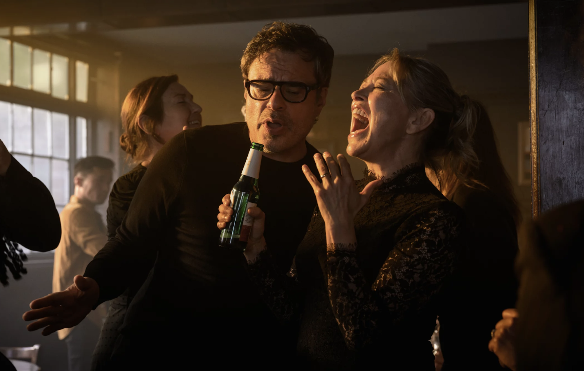 A middle aged man and woman stand in a crowded room singing enthusiastically into a beer bottle.