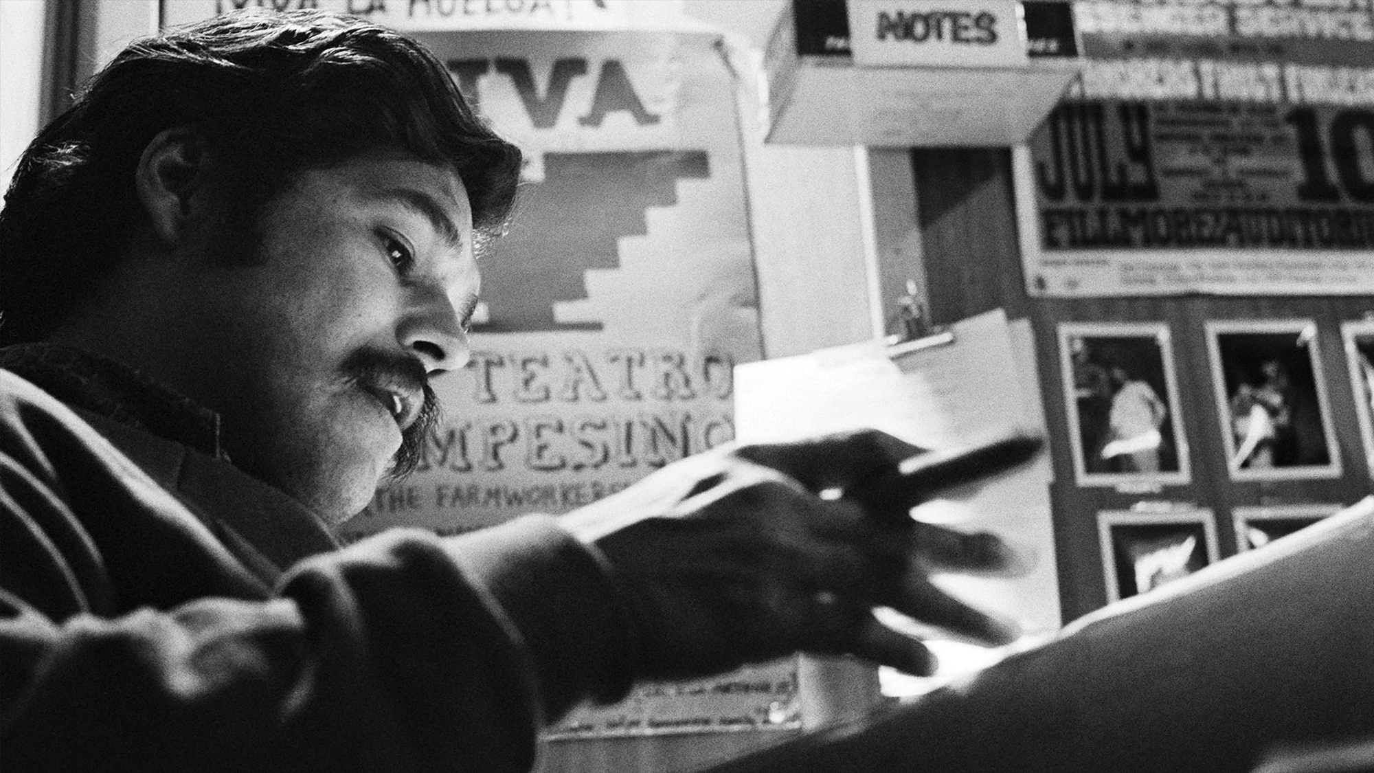 A Latino man, seen from a low angle, holds a pen and goes through papers against a wall background with theatre advertisements and photographs.
