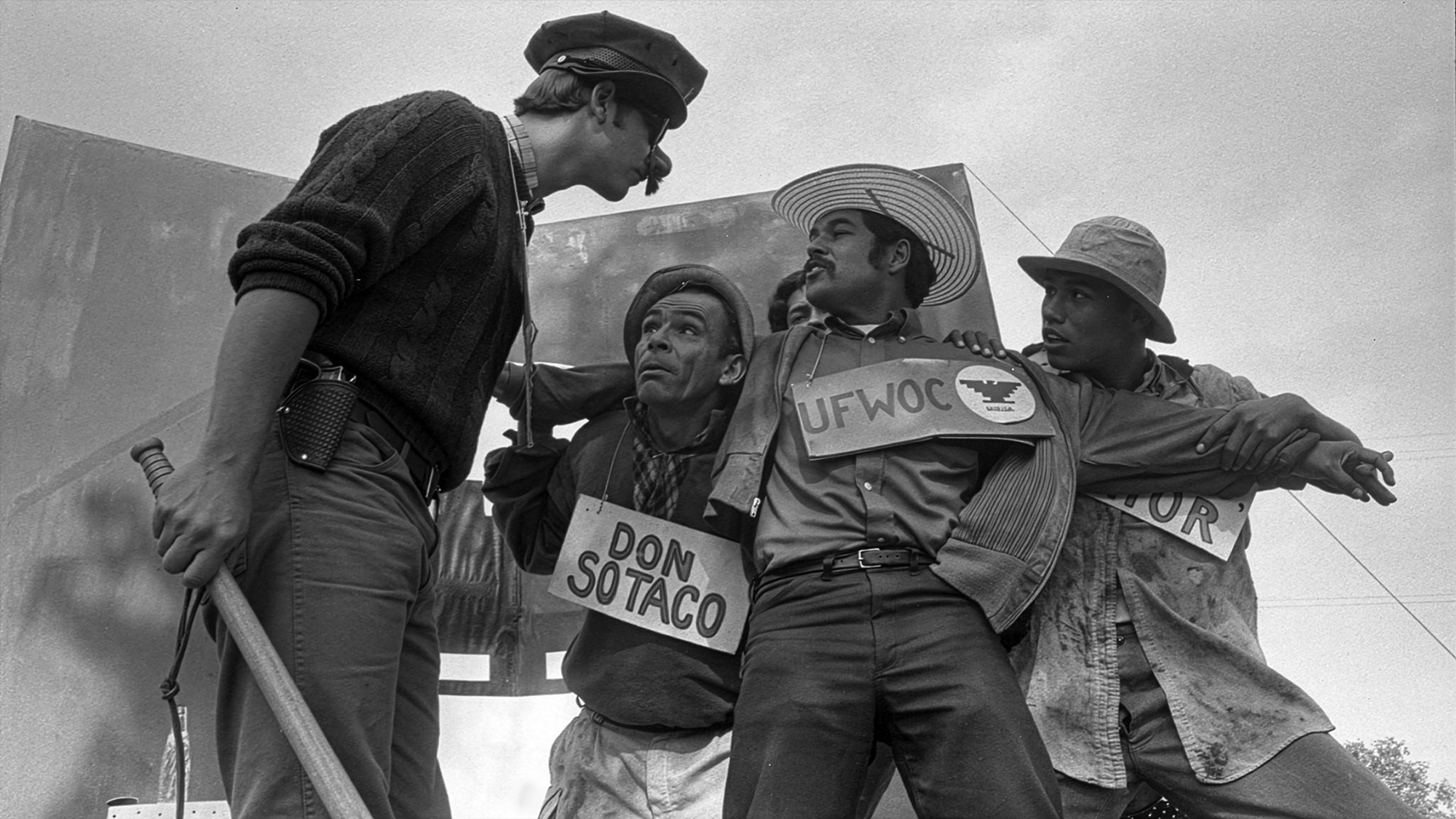 Four men, outdoors and seen from below, act out a scene dressed in simple clothes and wearing signs around their neck to indicate which group they belong to.