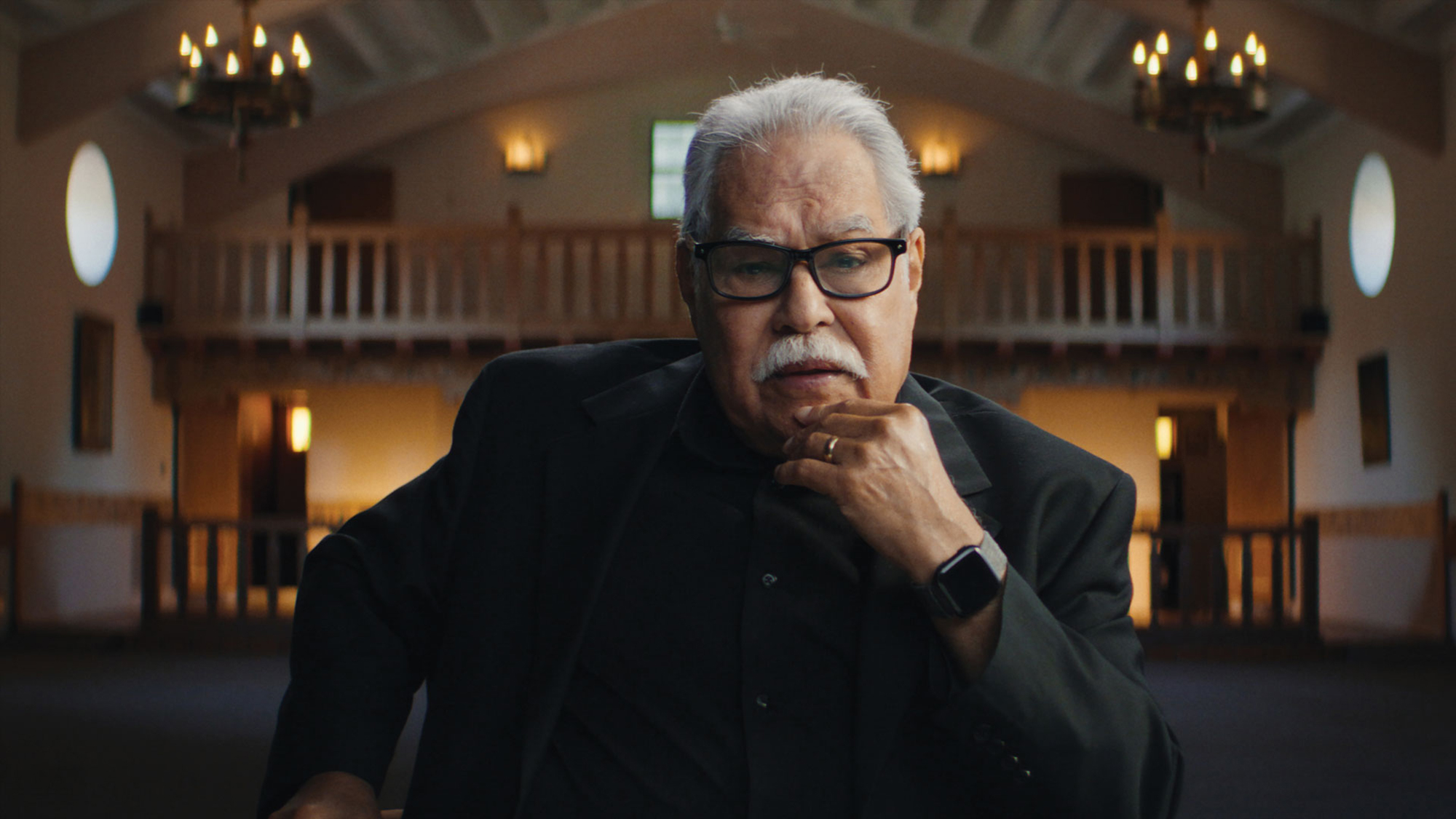 An older latino man in a Black jacket and glasses sits inside a high-ceiling church-like building, with chandeliers and wooden features