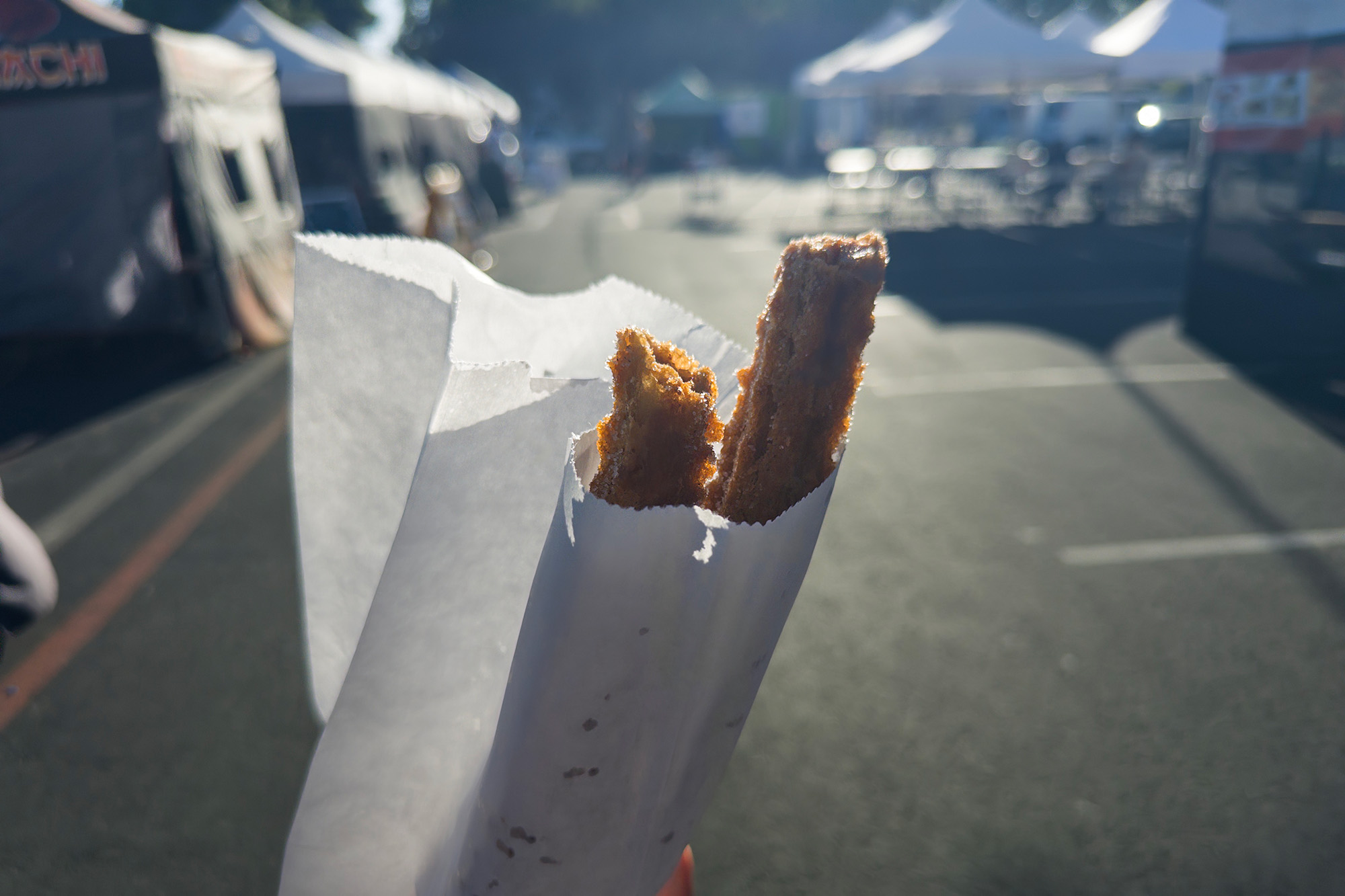 Two churros in a white paper bag, with food tents at a street market in the background.