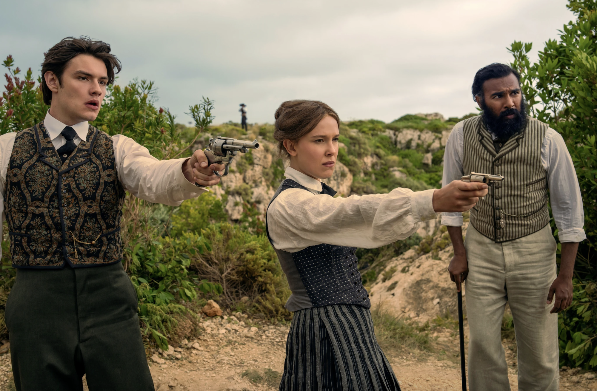 A woman in Victorian period costume stands in the desert pointing a small revolver. To her right is a white man doing the same. To her left, a man of Indian descent leaning on a walking stick.
