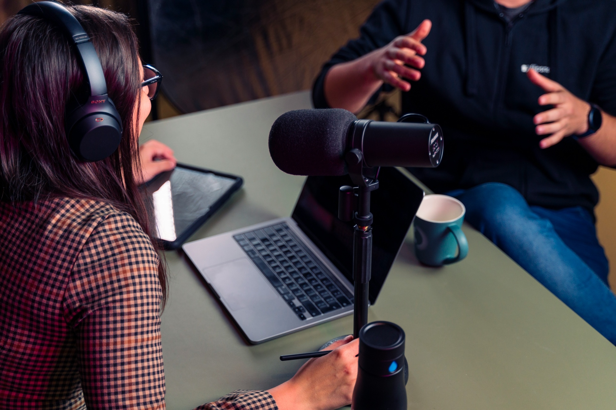 A young woman wearing headphones sits behind an open laptop and microphone and listens closely to a young man talking expressively opposite her.
