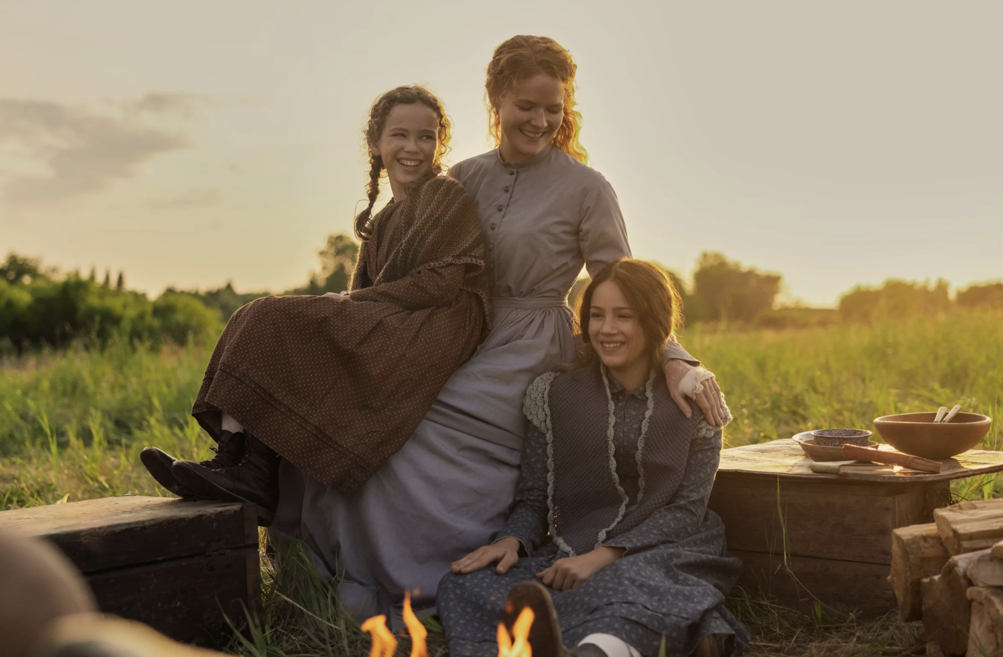 a white woman sits with her arms around her two daughters, with a backdrop of beautiful countryside. They are wearing pioneer-era dresses.