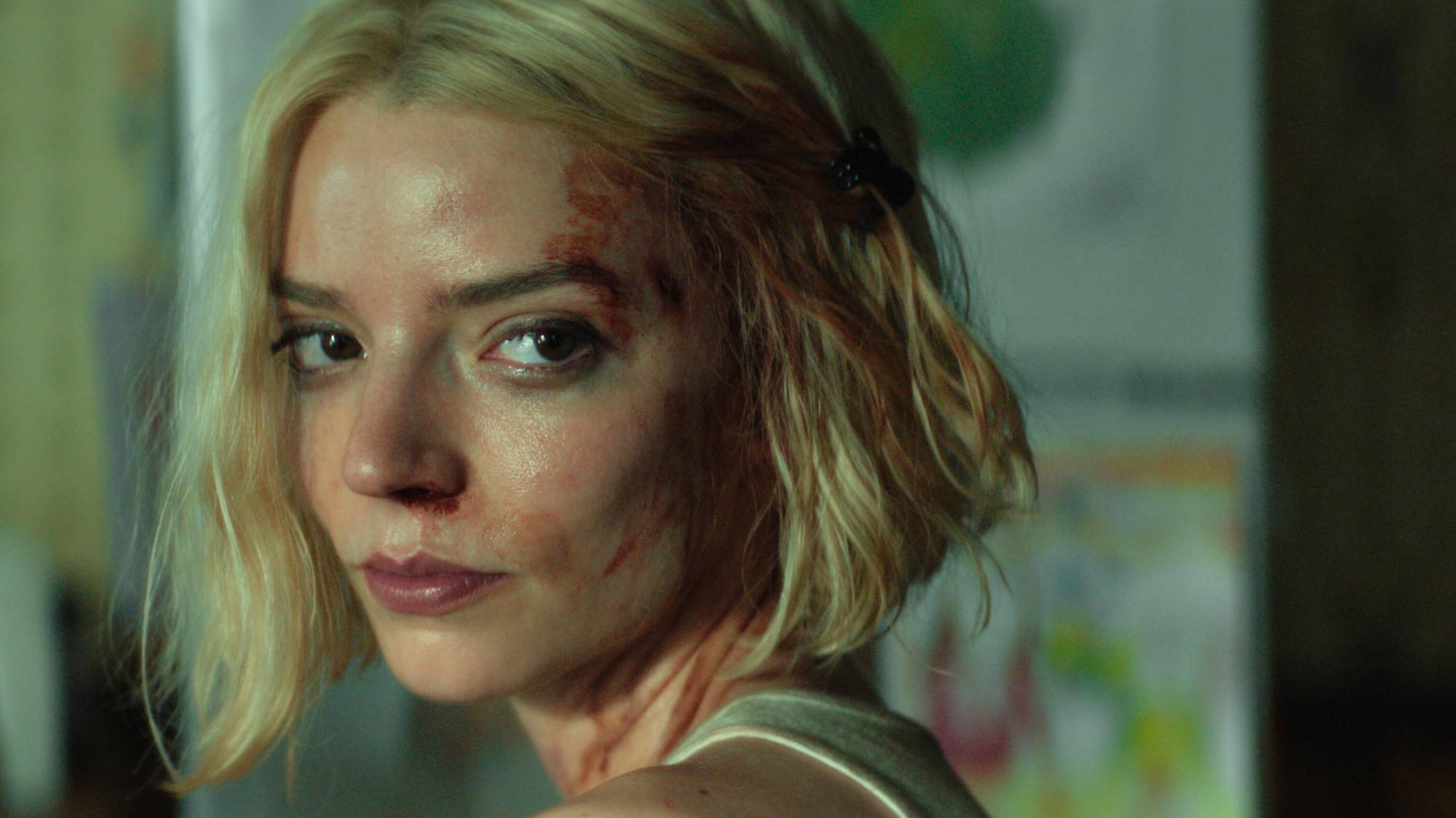 A young white woman with platinum blond hair looks over her shoulder, sweaty and disheveled with blood smeared on her face.