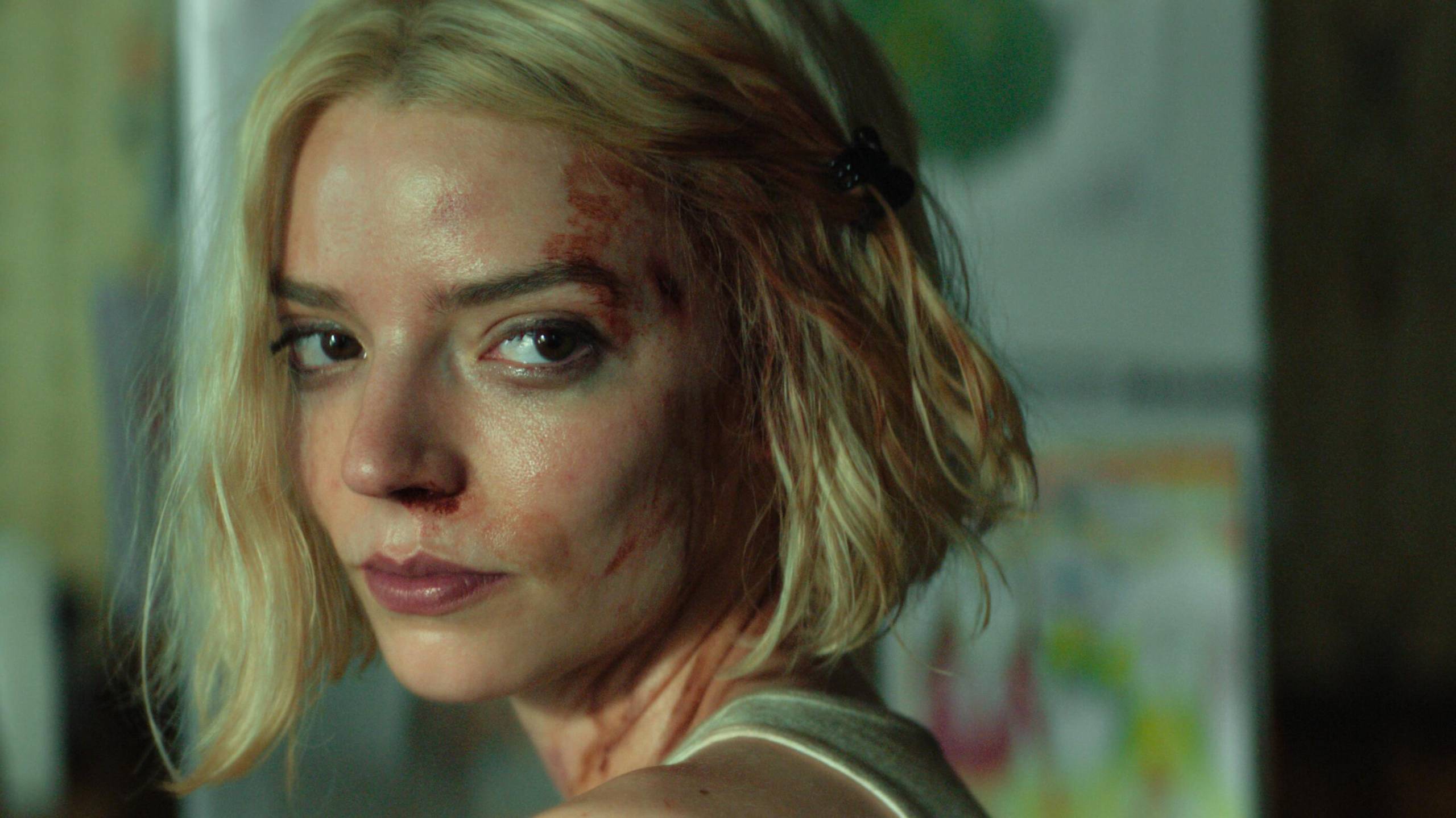 A young white woman with platinum blond hair looks over her shoulder, sweaty and disheveled with blood smeared on her face.