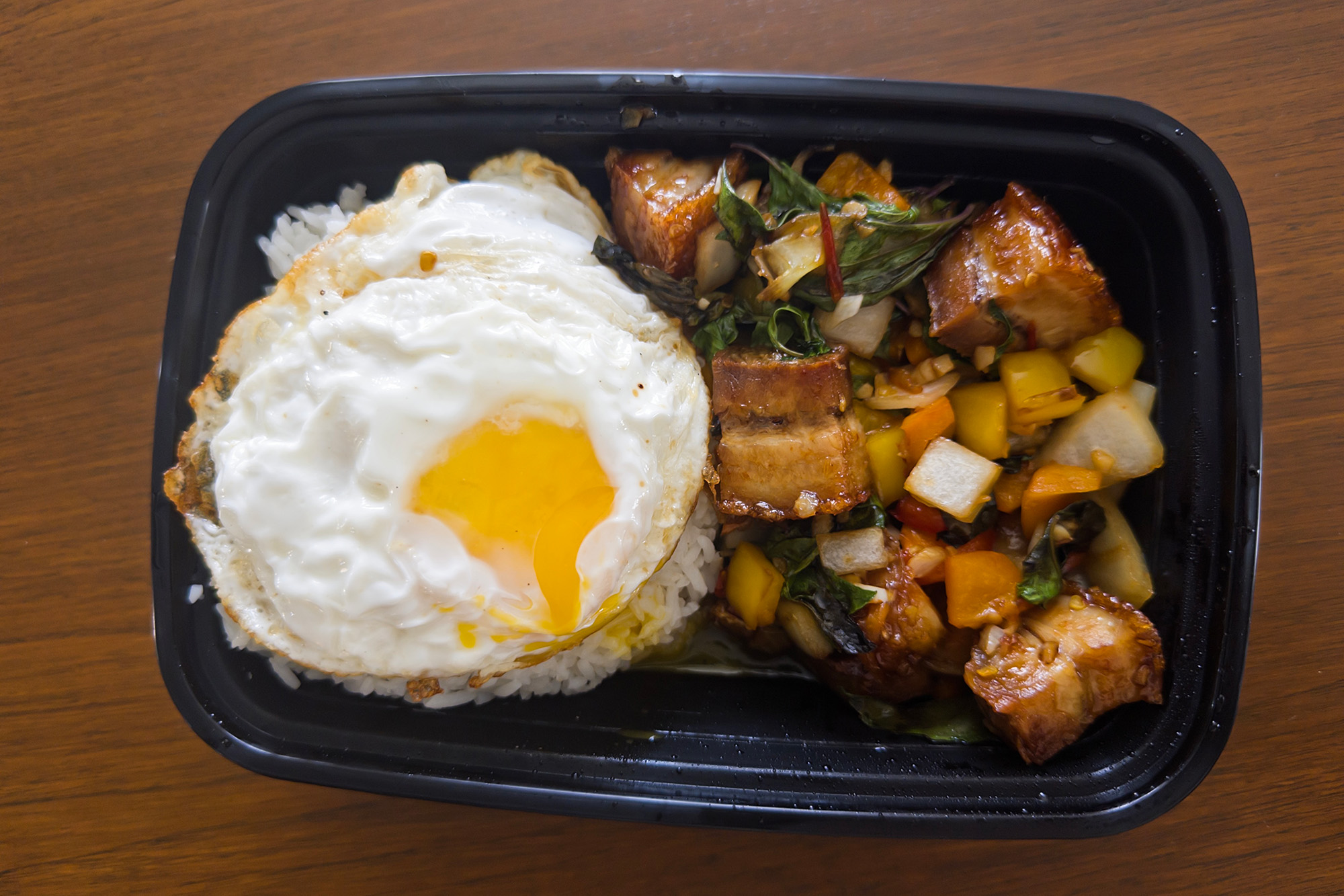 Takeout container with cubes of fried pork belly, basil, bell peppers and a fried egg.