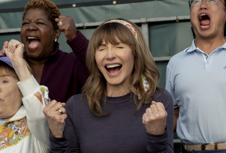 An attractive senior woman cheers with clenched fists in the stands of a sporting event. A Black woman and an Asian man stand slightly behind her also cheering enthusiastically.