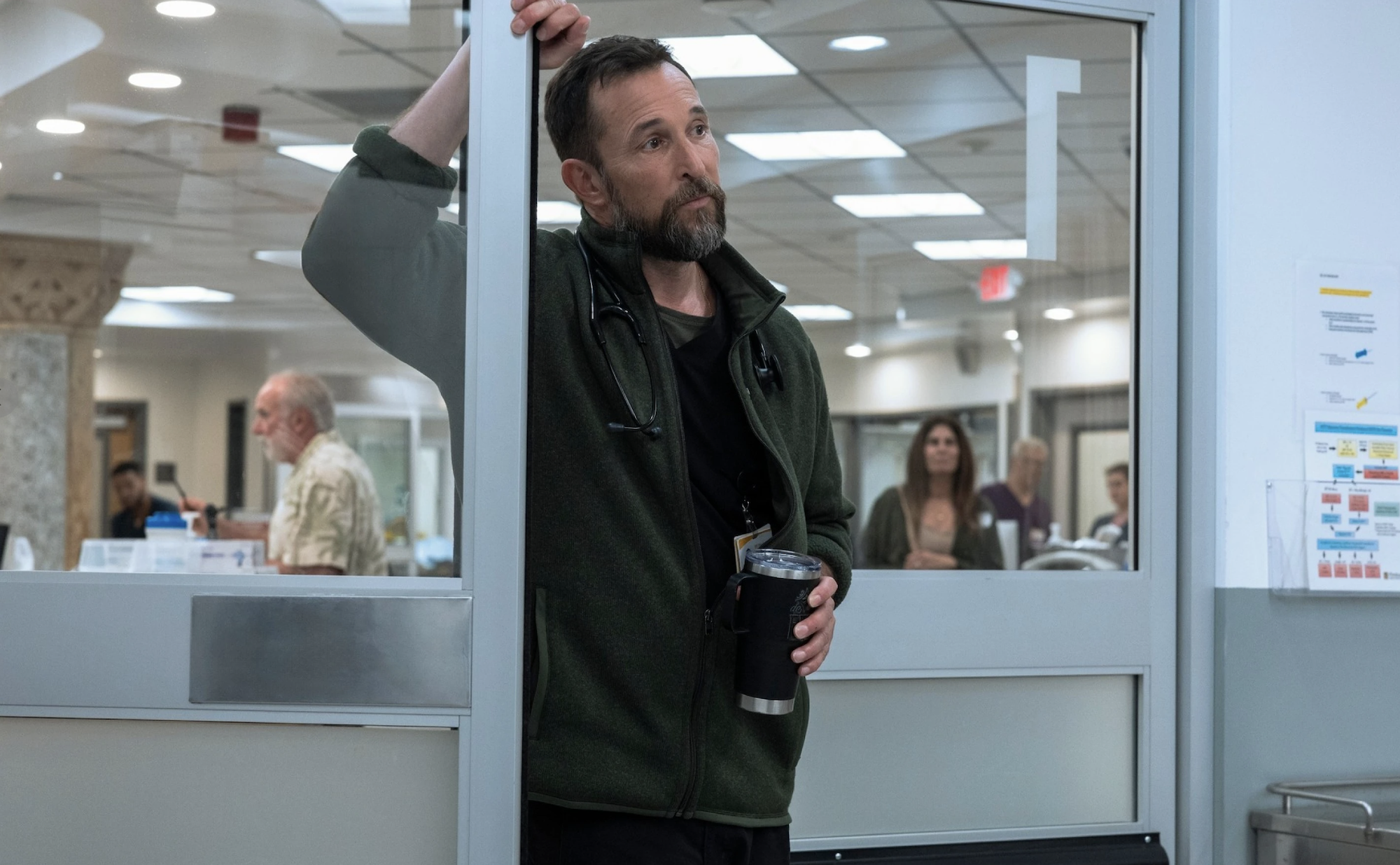 A bearded white man with a concerned expression stands in a glass doorway, clutching a large cup of coffee. He is wearing black pants, a black t-shirt and a fleece jacket. A stethoscope hangs around his neck.