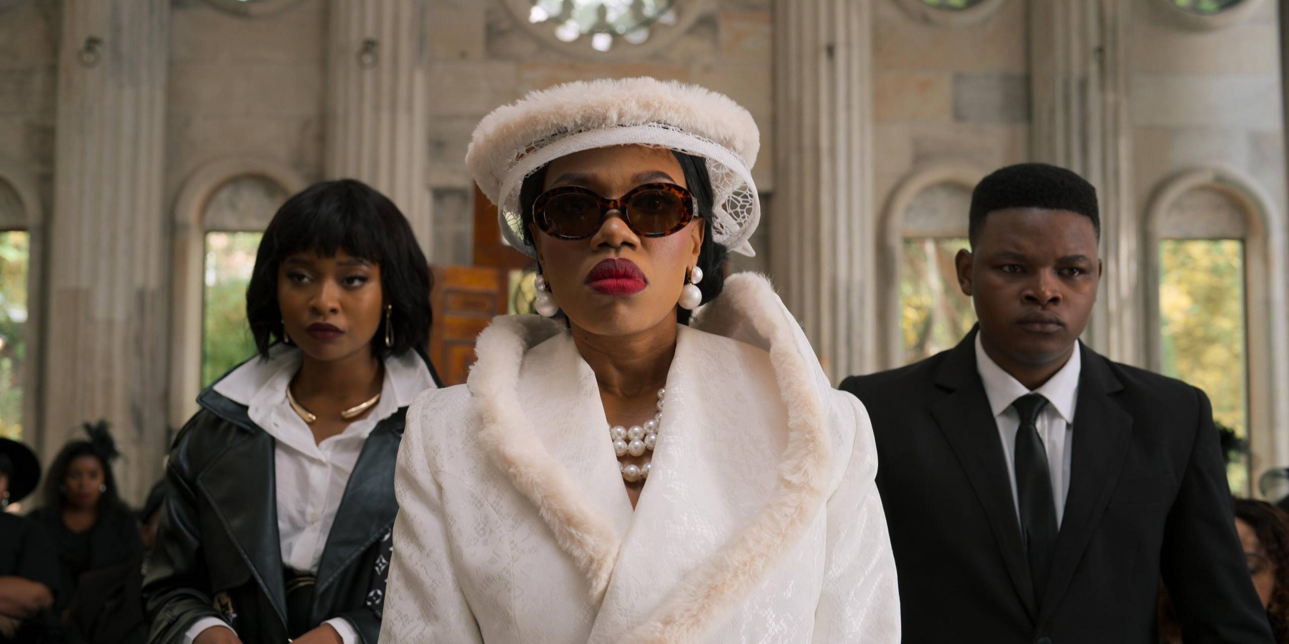 A well-groomed Black woman wearing a white suit and hat with dark sunglasses, walks into a palatial white room, with a Black man and woman either side of her, both wearing dark suits.