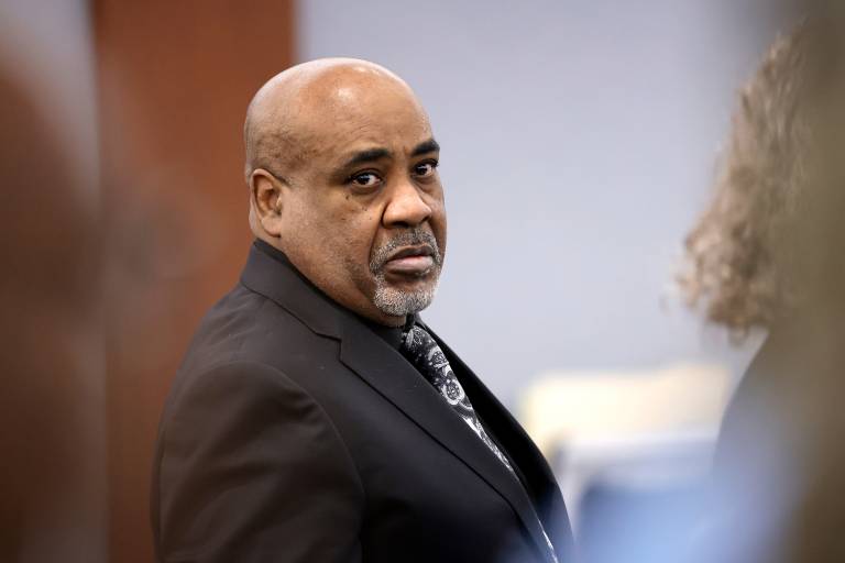 A middle aged Black man in a dark suit stands in a courtroom, looking to the right