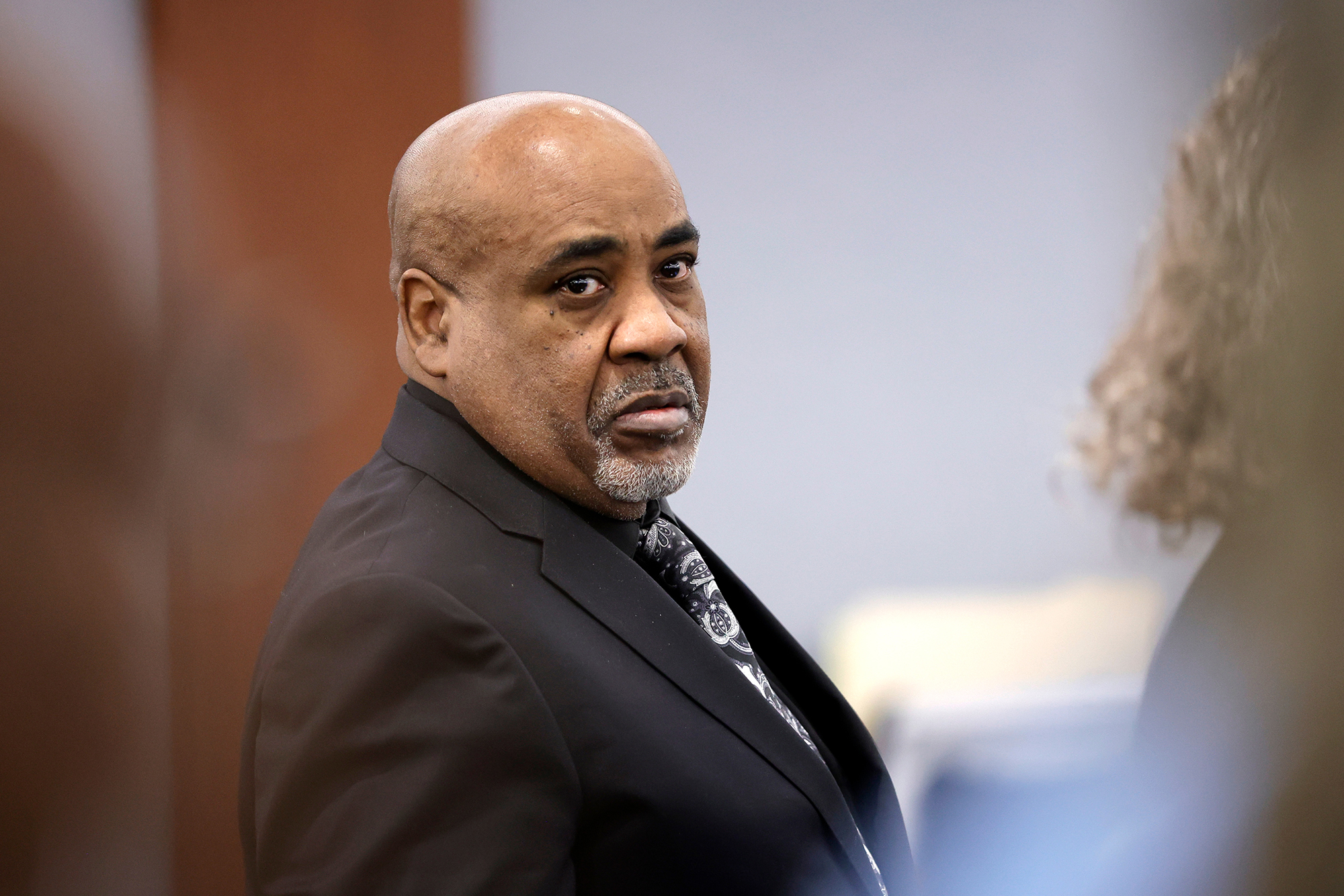 A middle aged Black man in a dark suit stands in a courtroom, looking to the right