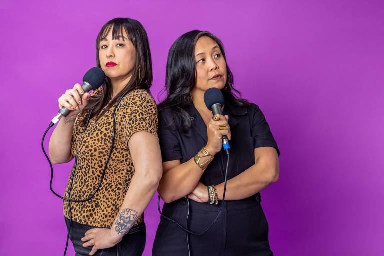 Two Asian American women stand before a purple backdrop, clutching microphones close to their faces. The woman on the left wears a leopard print t-shirt and black jeans; the one of the right wears a black jumpsuit.