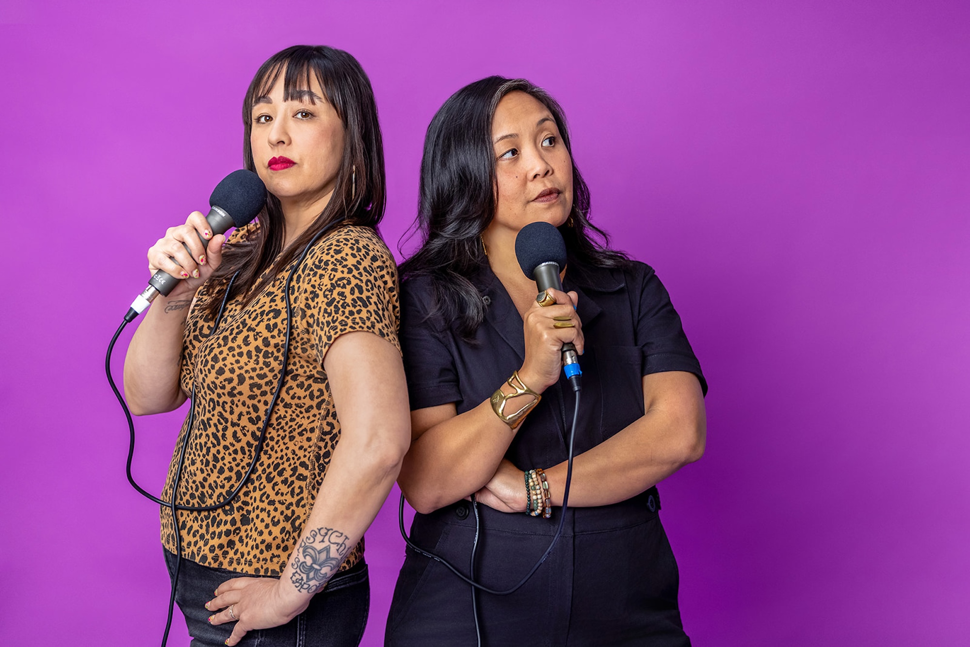 Two Asian American women stand before a purple backdrop, clutching microphones close to their faces. The woman on the left wears a leopard print t-shirt and black jeans; the one of the right wears a black jumpsuit.