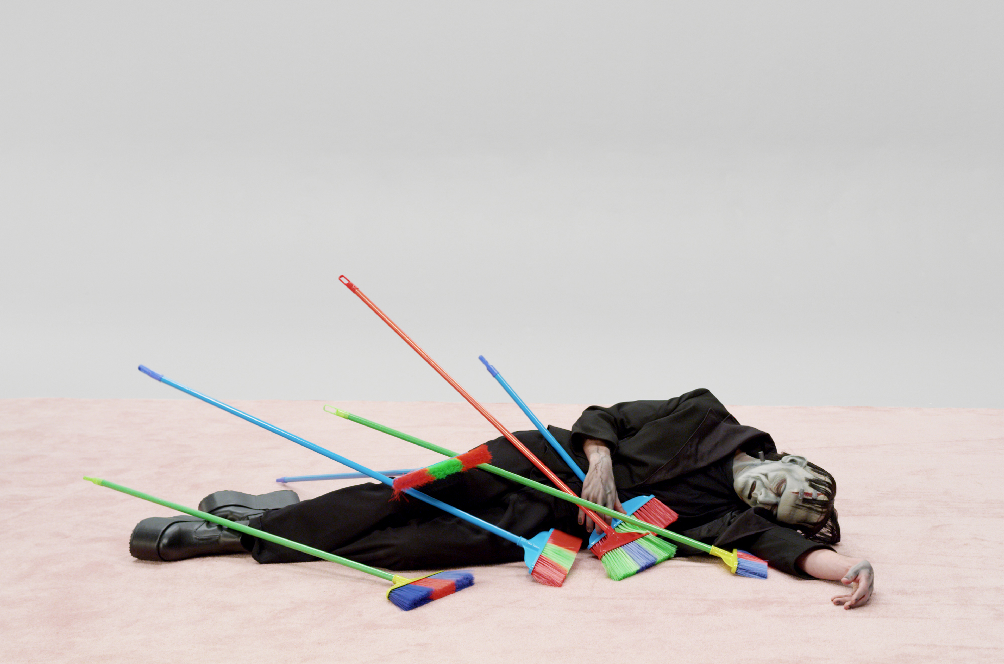 person in Frankenstein monster costume lays on ground with colorful plastic brooms