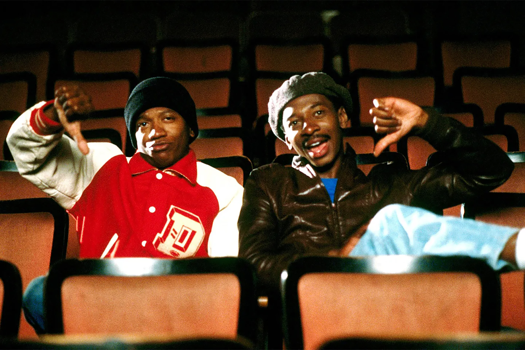 Two young Black men sit in an otherwise empty movie theater, both showing a thumbs-down hand signal