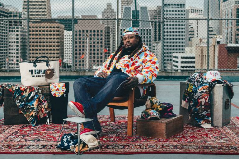 A Black man in glasses, backwards cap, jeans and colorful patterned jacket sits in a chair on an oriental rug with a backdrop of chain-link fencing and the San Francisco skyline