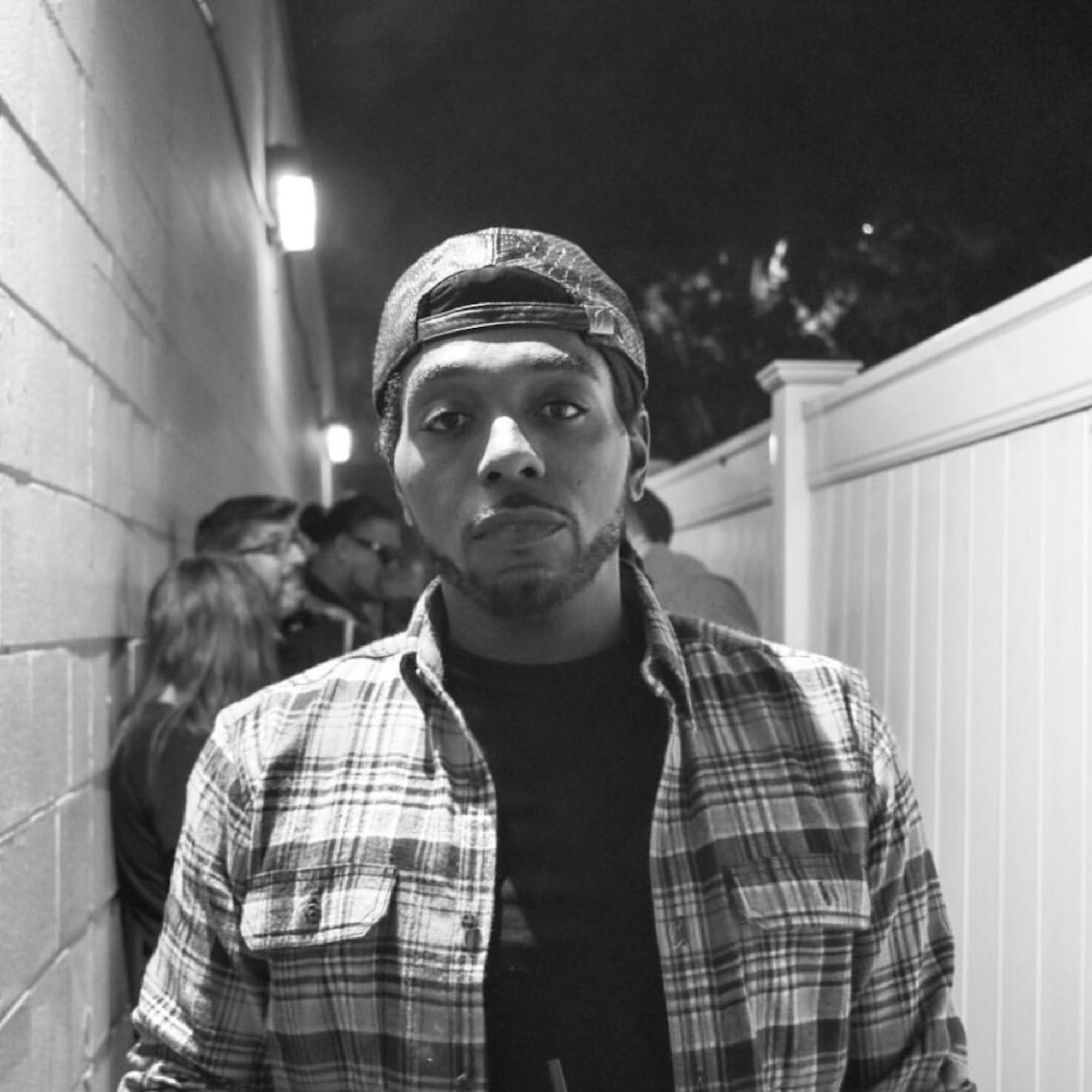A black and white photo of an African American man in a backwards hat and plaid shirt.