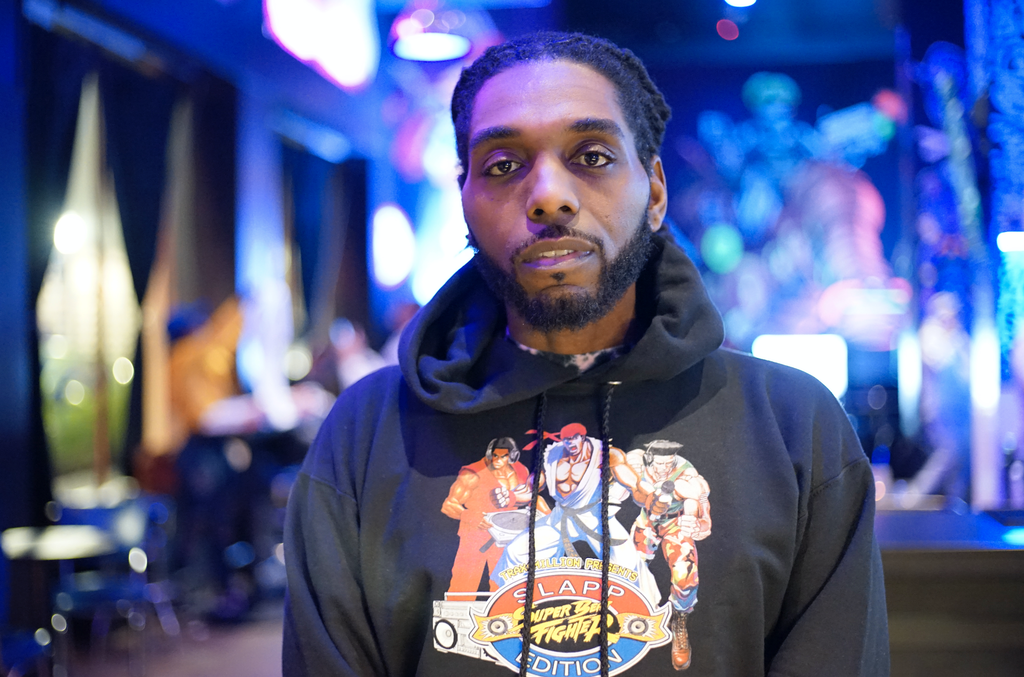 An African American man facing forward, looking at the camera as he wears a black hoodie adorned with animated characters.