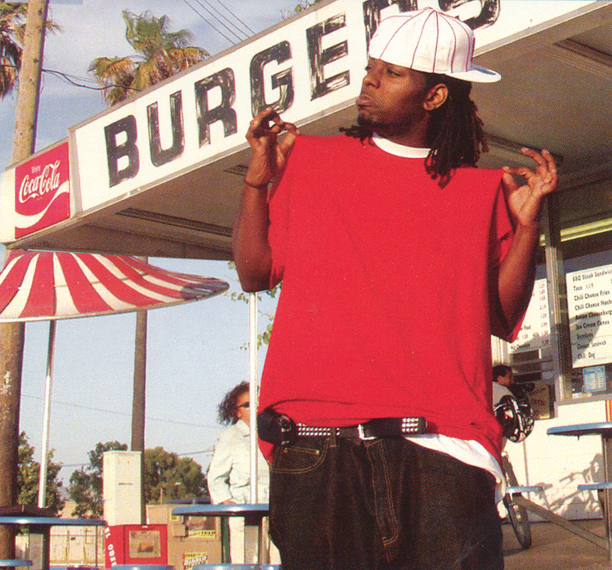 A young Black man in an oversized red T-shirt, backwards white striped baseball cap, studded belt and large jeans, in front of a hamburger stand