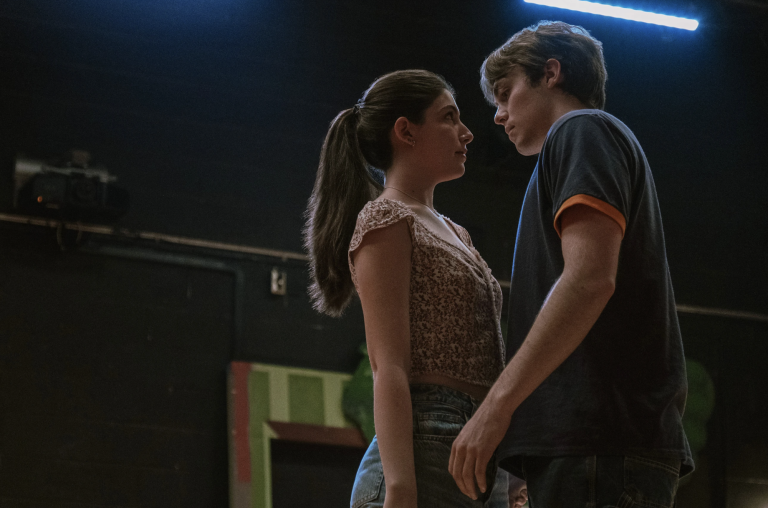 A teenage girl and boy stand close, face to face, in a dark studio.