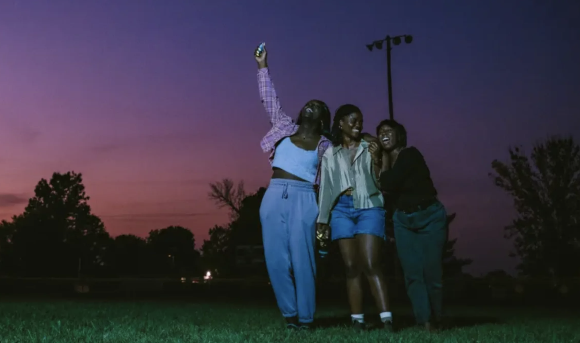 Three young Black women stand close together in a field at sunset, smiling and cheering, the sky purple behind them.
