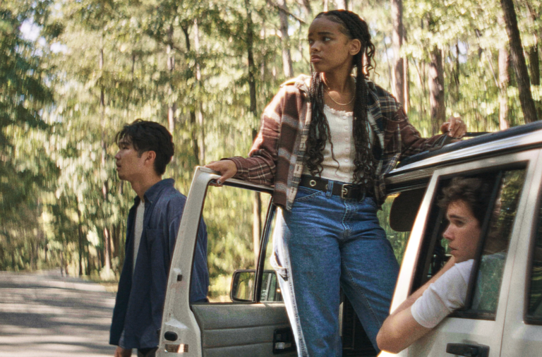 On a deserted road surrounded by forest, a white SUV is pulled over. A young Asian man stands at its side, a young Black woman stands in its front doorway and a young white man sits in the back with his elbow protruding from the open window. They are all dressed casually.