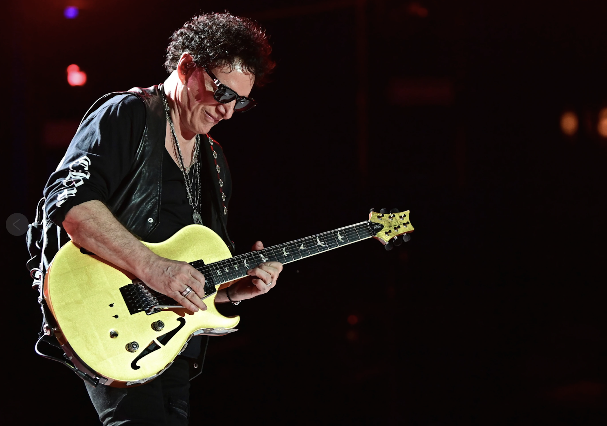 A senior white man with brown hair, dressed in all black and sunglasses plays a yellow electric guitar on stage.