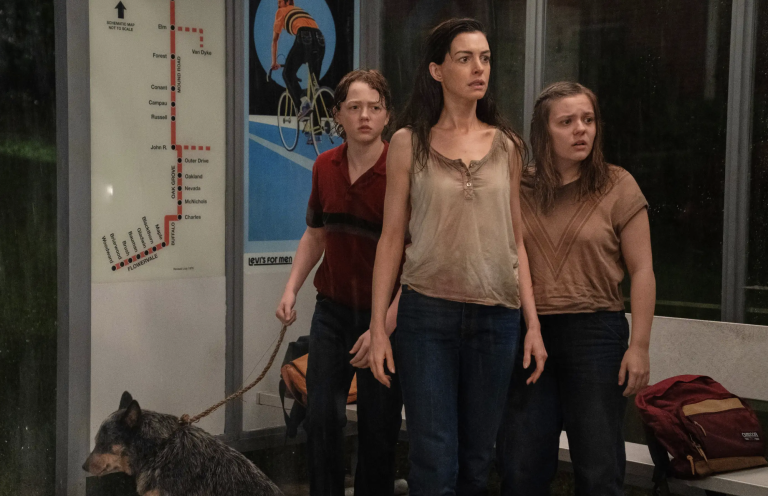 A bedraggled middle aged white woman with long dark hair stands between a young boy and teenage girl, looking worried. The boy has a dog on a leash. They are standing inside a subway car.
