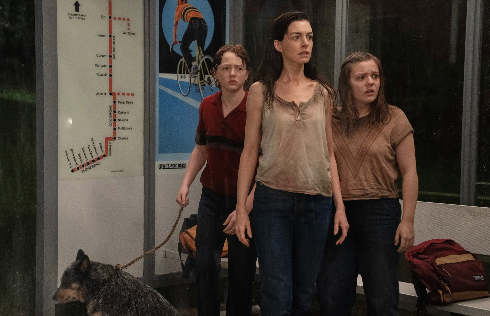 A bedraggled middle aged white woman with long dark hair stands between a young boy and teenage girl, looking worried. The boy has a dog on a leash. They are standing inside a subway car.