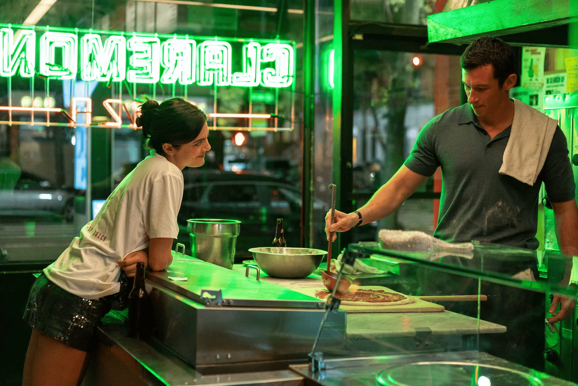 A woman leans over the counter, looking at a man making pizza.