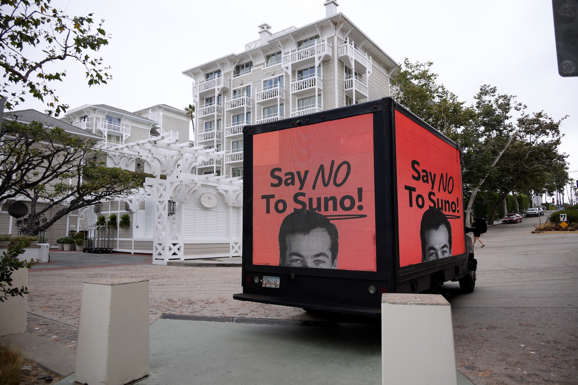 A black truck displaying electronic screens with the message SAY NO TO SUNO drives past a white building.