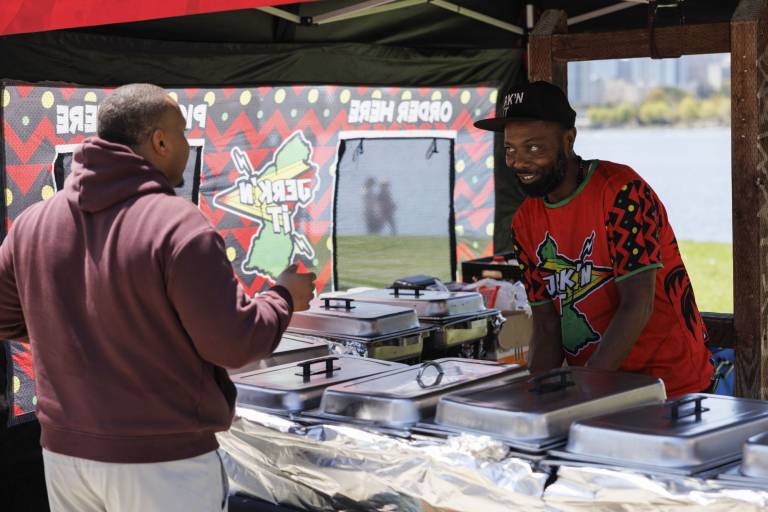 A chef in a baseball cap talks to a customer buying food from his food tent. A lake is visible in the background.