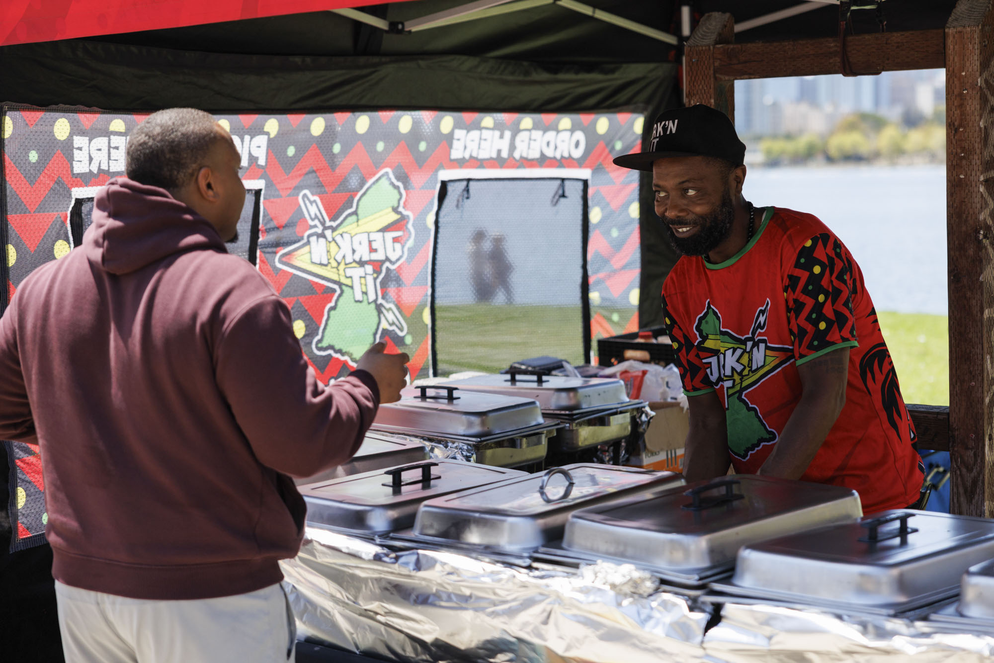 A chef in a baseball cap talks to a customer buying food from his food tent. A lake is visible in the background.