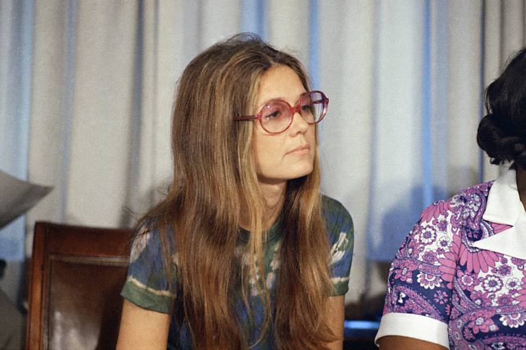 A young woman with long dirty blonde hair parted in the middle in a green top with glasses looks off to the right