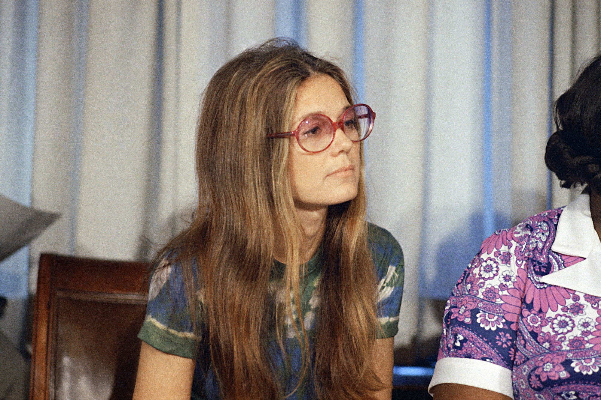 A young woman with long dirty blonde hair parted in the middle in a green top with glasses looks off to the right