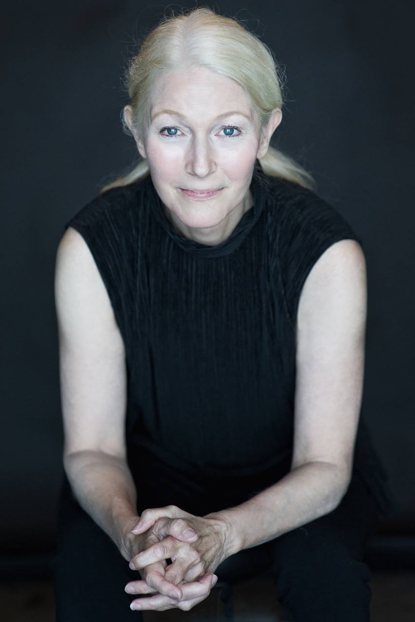 white woman with light hair leans forward with slight smile, hands clasped